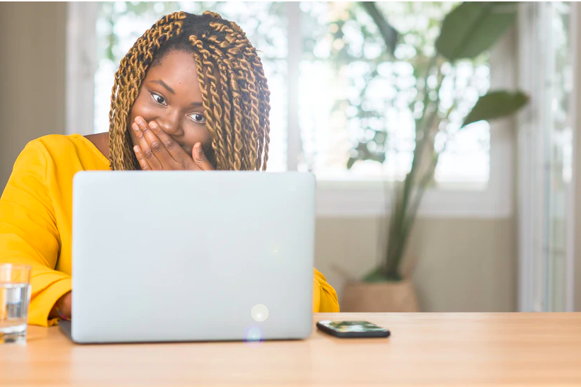 woman in yellow holding her hand over her mouth while looking at a laptop