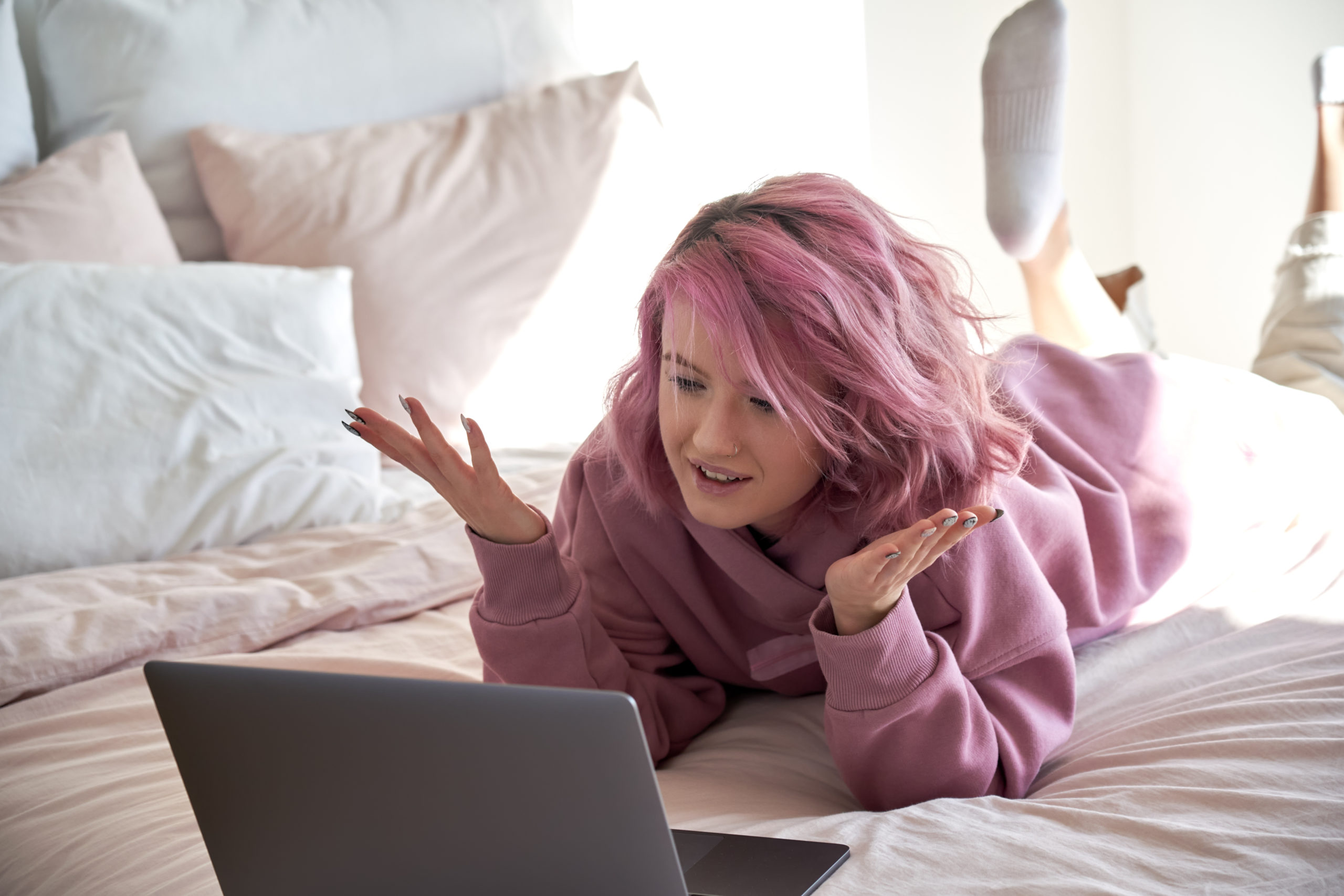 woman with pink hair wears hoodie video calling looking at laptop lying in bed.