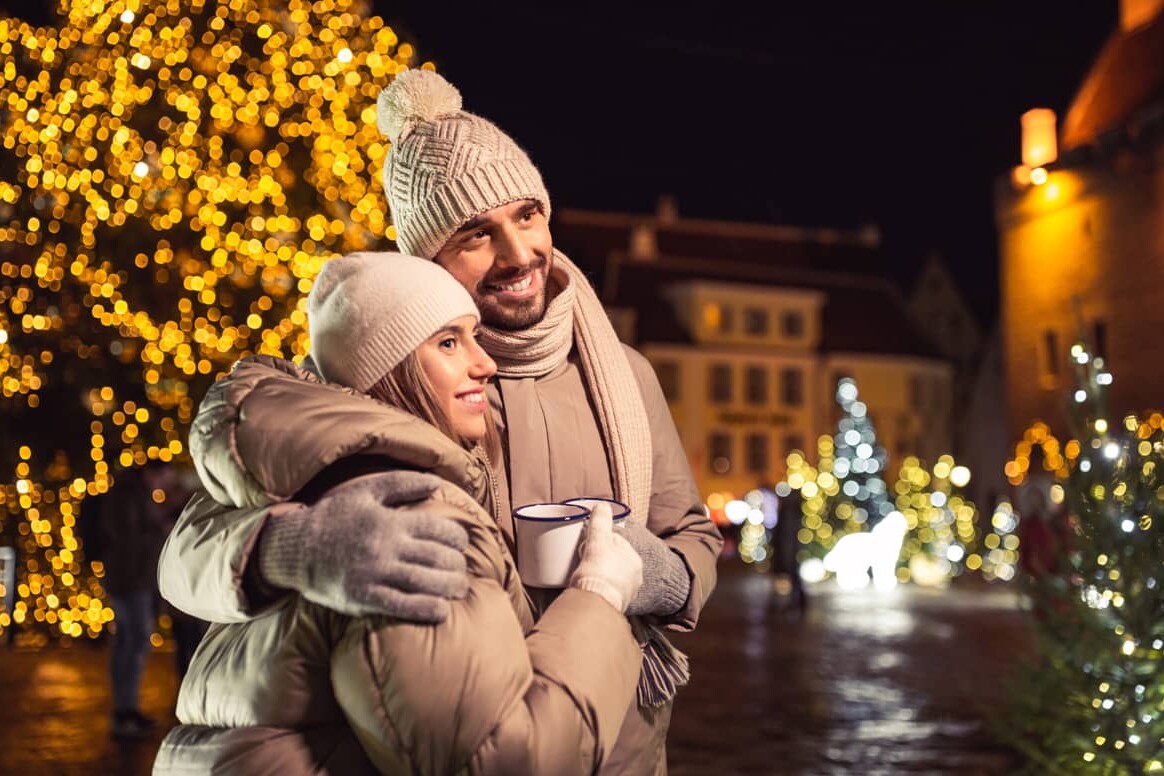 couple wearing winter clothes and hugging and holding mugs looking at holiday lights