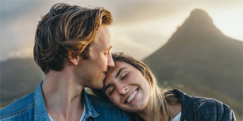 Close-up of smiling beautiful young girlfriend with eyes closed resting head on boyfriend' shoulder against mountain at sunset